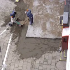 Pracownicy układają materiał na stary chodnik betonowy. Wózek i łopaty wskazują na prace budowlane.