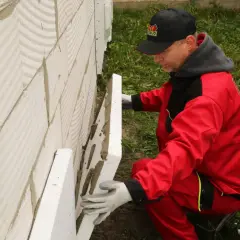 Budowlaniec w czerwonym stroju montuje styropian na ścianie z bloczków. To kluczowy etap prac, by zapewnić odpowiednią izolację, a pytanie brzmi: ile styropianu na strop betonowy?
