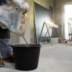 Robotnik wsypuje suchą mieszankę do wiadra, przygotowując beton na styropianie. Kluczowa jest minimalna grubość wylewki betonowej na styropianie.
