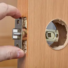 Dłoń wsuwa metalowy mechanizm zamka do wyciętego otworu w drewnianych drzwiach.