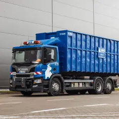 Niebieska śmieciarka z kontenerem na gruz betonowy, z logo BYŚ Recycling, stoi przed szarym budynkiem.
