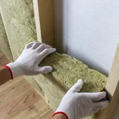 Ręce w białych rękawiczkach dopasowują wełnę mineralną do drewnianej konstrukcji. Do cięcia wełny mineralnej użyj specjalnego noża.