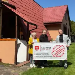 Trzy osoby trzymają baner "Budynek zalegalizowany" przed domem. Dowiedz się, jak zrobić odbiór domu po 20 latach.