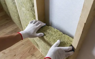 Ręce w białych rękawiczkach dopasowują wełnę mineralną do drewnianej konstrukcji. Do cięcia wełny mineralnej użyj specjalnego noża.