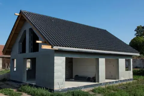 Dom w stanie surowym otwartym, z ciemnym dachem i betonowymi ścianami, czeka na dalsze prace budowlane.