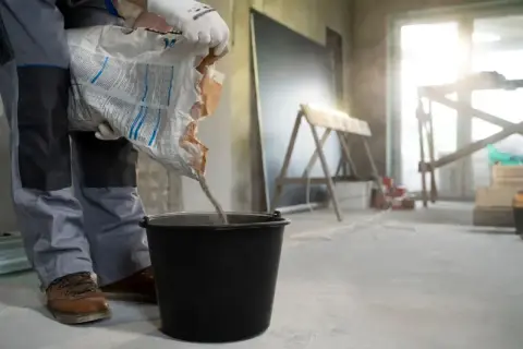 Robotnik wsypuje suchą mieszankę do wiadra, przygotowując beton na styropianie. Kluczowa jest minimalna grubość wylewki betonowej na styropianie.