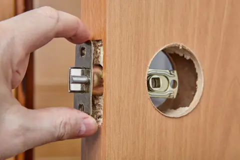 Dłoń wsuwa metalowy mechanizm zamka do wyciętego otworu w drewnianych drzwiach.