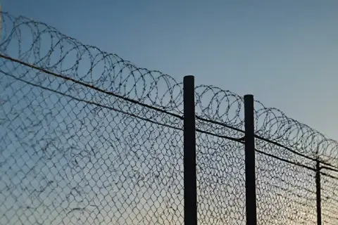 Ogrodzenie z siatki z nawiniętym drutem kolczastym na słupkach, na tle błękitnego nieba.