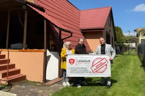 Trzy osoby trzymają baner "Budynek zalegalizowany" przed domem. Dowiedz się, jak zrobić odbiór domu po 20 latach.