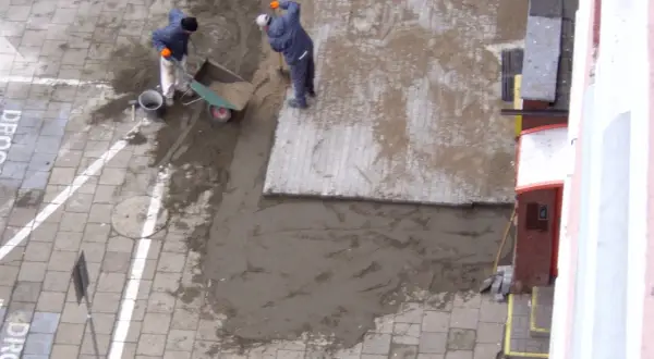 Pracownicy układają materiał na stary chodnik betonowy. Wózek i łopaty wskazują na prace budowlane.