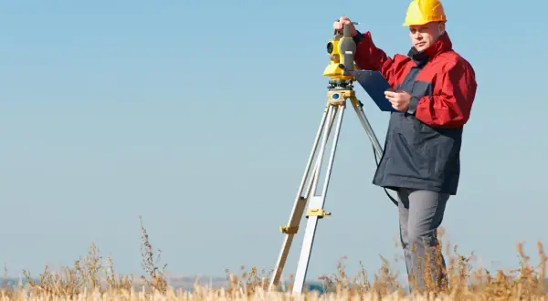 Geodeta w żółtym kasku pracuje przy niwelatorze. Zastanawiasz się, ile kosztuje geodeta przy budowie domu?