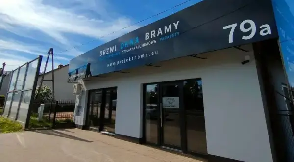 Nowoczesny budynek z dużymi oknami i drzwiami, z banerem "DRZWI OKNA BRAMY".