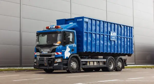 Niebieska śmieciarka z kontenerem na gruz betonowy, z logo BYŚ Recycling, stoi przed szarym budynkiem.