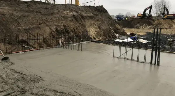 Świeża wylewka betonowa na zewnątrz, zbrojenie i koparki w tle.