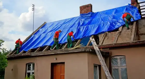 Pracownicy w zielonych ogrodniczkach i czerwonych koszulkach zabezpieczają dach niebieską plandeką podczas przebudowy domu.