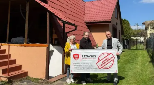 Trzy osoby trzymają baner "Budynek zalegalizowany" przed domem. Dowiedz się, jak zrobić odbiór domu po 20 latach.