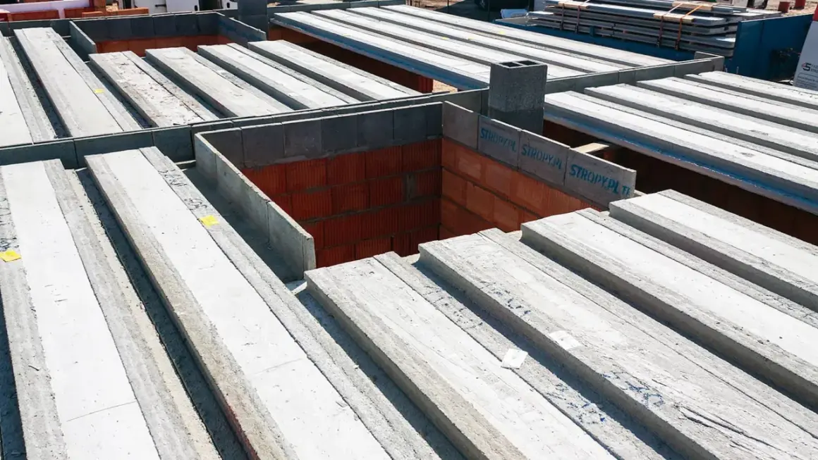 Budowa domu w toku: betonowe belki stropowe czekają na montaż, tworząc solidny strop nad ceglanymi ścianami.