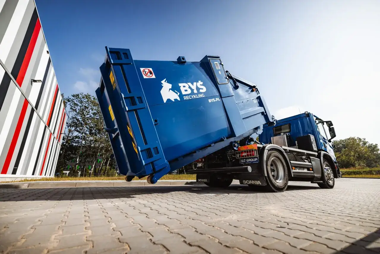 Niebieska śmieciarka z logo BYŚ Recykling podnosi kontener, gotowa do odbioru gruzu betonowego.